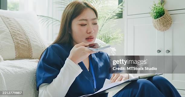 young asian female nurse in blue scrubs using voice recognition technology on phone while reviewing patient care documents on a clipboard in living room at home. nurse sustainable lifestyle concept. - speech recognition stock pictures, royalty-free photos & images