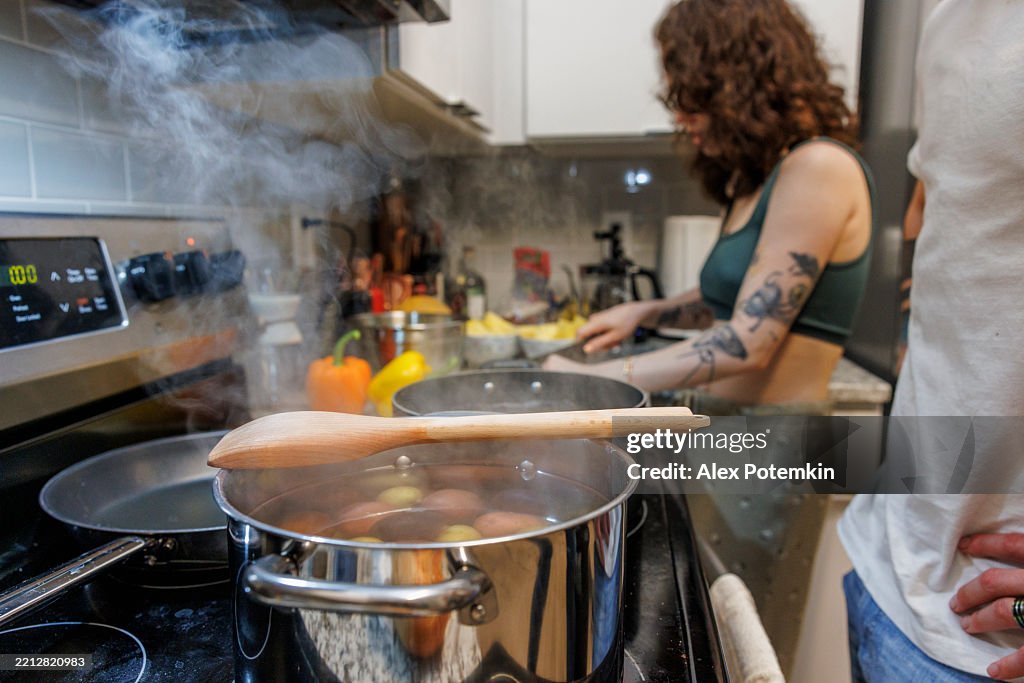 Bouillon de pommes de terre bouilli dans une casserole fumante avec une cuillère en bois. Un homme méconnaissable avec des mains sur les hanches attendant près du poêle. Femme brune avec tatouage coupant des légumes sur fond flou.