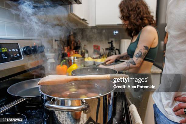 boiled potato broth in steamy pot with wooden spoon. unrecognizable man with hands on hips waiting by the stove. brunette woman with tattoo cutting vegetables on blurred background. - gekookte aardappel stockfoto's en -beelden