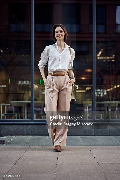 stylish young woman with confident stride walking in front of city glass building - figura intera foto e immagini stock