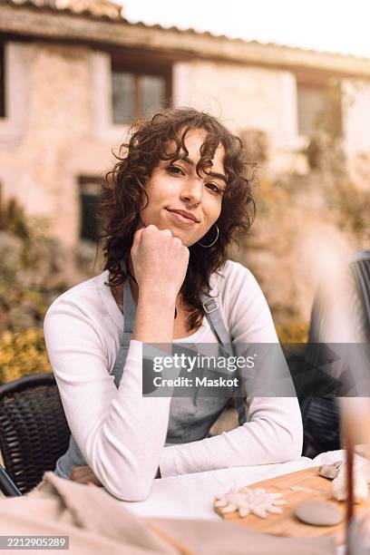 portrait of smiling woman with curly hair sitting at table - hand on chin stock pictures, royalty-free photos & images