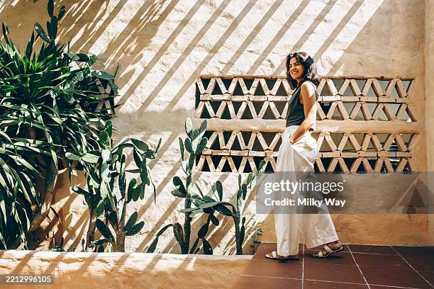 latina woman in white pants and green top walks next to a textured wall with cactus plants. sunlight casts long shadows, creating a geometric pattern. - educational subject stock pictures, royalty-free photos & images