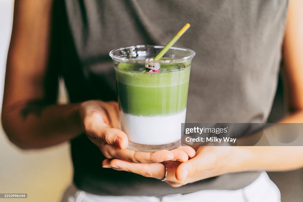 Close-up of layered ice matcha drink in a stylish glass with a yellow straw and edible flower garnish, held by both hands.