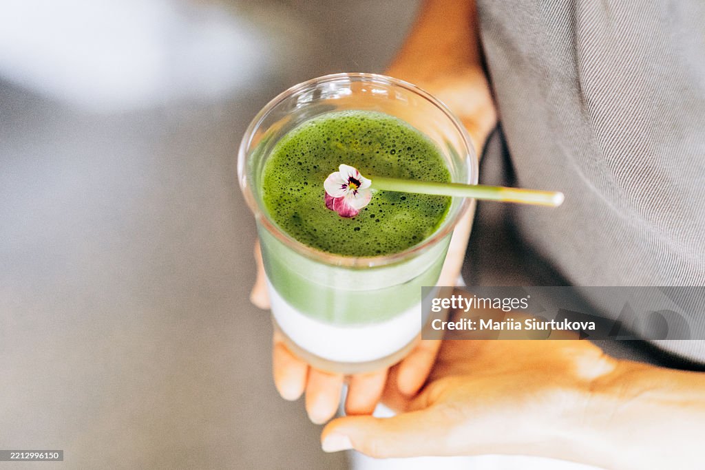 Close-up of hands holding a matcha latte topped with foam and an edible flower. Layered green and white drink in a clear glass with a gold spoon. Casual summer outfit visible.