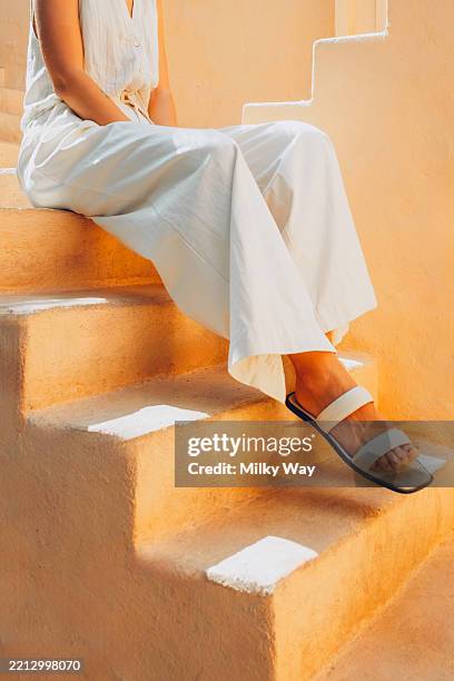 a woman dressed in loose white clothing sits on beige stairs. only her legs and sandals are visible in the frame. warm natural lighting. - ambiente atmosférico fotografías e imágenes de stock