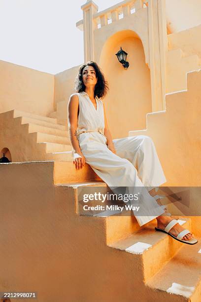 latina woman sits on beige stairway in sleeveless white outfit. she looks up slightly and smiles. mediterranean-style architecture with warm sunlight. - temas de mujeres fotografías e imágenes de stock