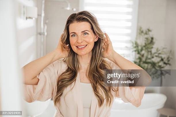 mujer cepillando el cabello con un peine mientras está de pie cerca del espejo en el baño. - mujeres de mediana edad fotografías e imágenes de stock