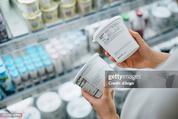 close-up shot of a woman shopping for fresh organic yoghurt in supermarket, reading and comparing the nutrition label and ingredients on the pot. routine grocery shopping. making healthier food choices. healthy eating lifestyle. consumer awareness concept - levend organisme stockfoto's en -beelden