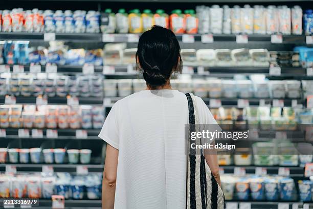 rear view of young asian woman doing grocery shopping, standing in front of a shelf full of fresh dairy products in the dairy aisle and thinking which product to choose from in a supermarket. concept of customer awareness - stormarknad bildbanksfoton och bilder