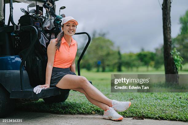 golfista femenina asiática mirando a la cámara relajándose por el carrito de golf durante el juego al aire libre - pies cruzados en los tobillos fotografías e imágenes de stock