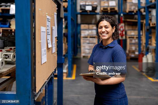 warehouse employee looking at her shifts on a bulletin board and smiling - trabalhador de armazém imagens e fotografias de stock