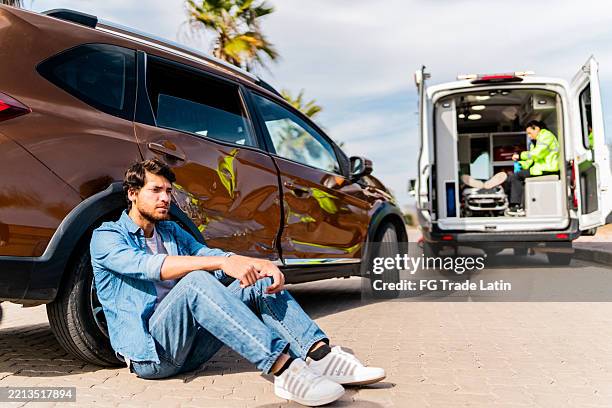mann sitzt nach autounfall auf dem boden mit krankenwagen im hintergrund - autounfall stock-fotos und bilder