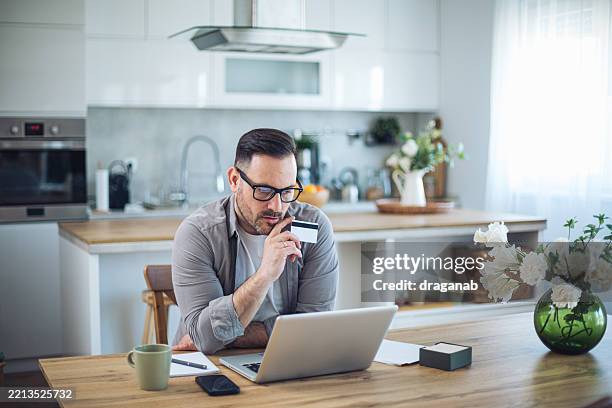 hombre adulto medio de compras en línea en cocina moderna - pago con tarjeta fotografías e imágenes de stock
