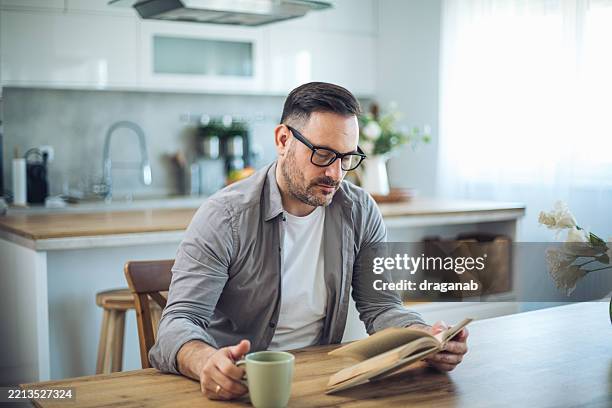 mid adult man reading a book and drinking coffee in his kitchen - reading glasses stock pictures, royalty-free photos & images