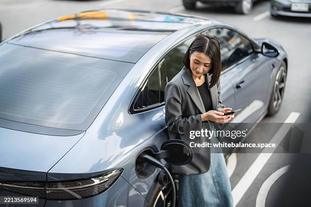 a confident businesswoman sitting in her ev car at the charging station, using a laptop while discussing work on her smartphone - charging stock pictures, royalty-free photos & images