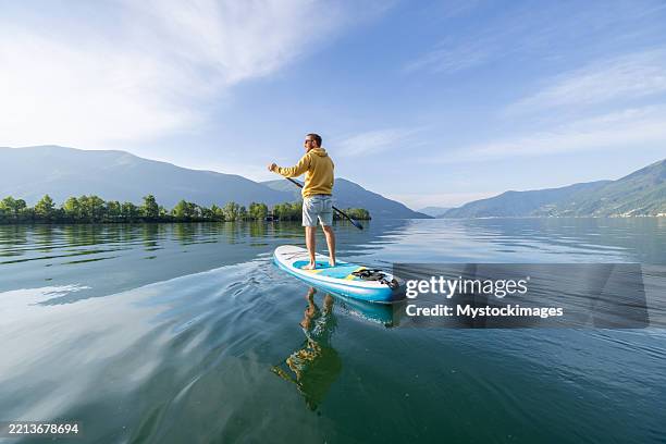 man paddleboarding on serene lake with mountain views - paddleboard stock pictures, royalty-free photos & images