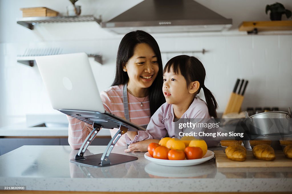 Una madre asiática ayuda a su pequeña hija a estudiar mientras está sentada en la cocina con su computadora portátil
