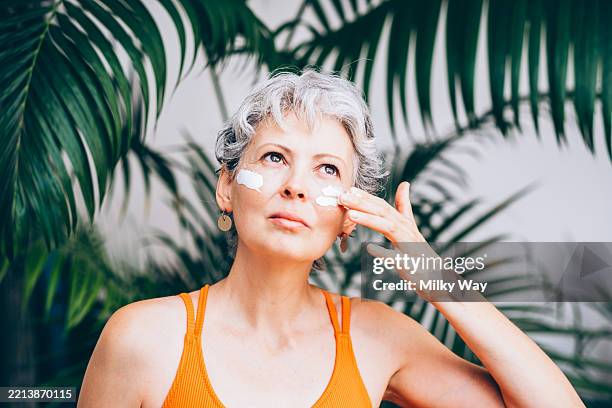 young woman with short grey curly hairstyle applies cream to her face. woman in an orange swimsuit takes care of skin during summer vacation in hot climate. lush palm leaves fill the tropical background. - sunscreen face stock pictures, royalty-free photos & images