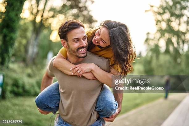 young man giving piggyback ride to his girlfriend in a park - piggyback stock pictures, royalty-free photos & images