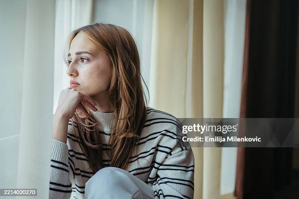 sad, depressed young woman sitting on the floor by the window. portrait of lonely, unhappy blond woman. - world mental health day stock pictures, royalty-free photos & images