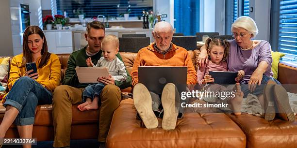 mehrgenerationenfamilie, die sich mit digitalen geräten auf einem gemütlichen sofa im warm beleuchteten wohnzimmer beschäftigt - websurfen stock-fotos und bilder