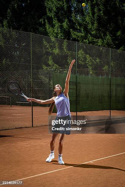 athletic female tennis player serving in purple tank top and navy skirt on clay court - green sweater vest stock pictures, royalty-free photos & images