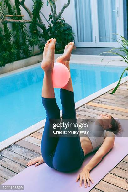 woman lying on yoga mat with legs raised, holding a pink exercise pilates ball between feet. poolside. - pelvic floor stock pictures, royalty-free photos & images