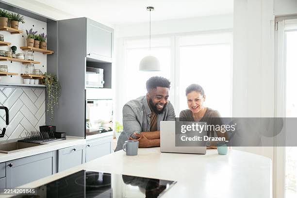 multiracial couple sits at a kitchen table working on a laptop, smiling and using internet - laptop kitchen table stock pictures, royalty-free photos & images