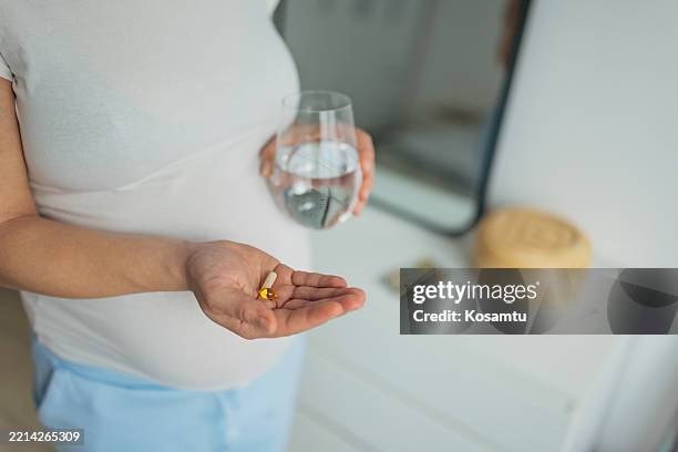 unrecognizable pregnant woman holding a glass of water, taking supplements for a healthy pregnancy while standing in her bedroom. prenatal vitamins - magnesium stock pictures, royalty-free photos & images