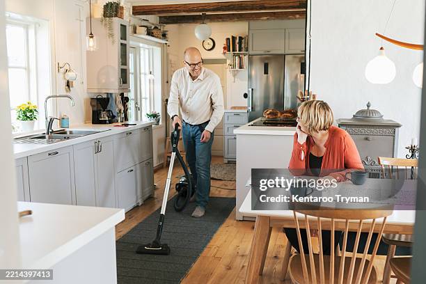 a man vacuuming the kitchen while his wife drinks coffee. - stofzuiger stockfoto's en -beelden