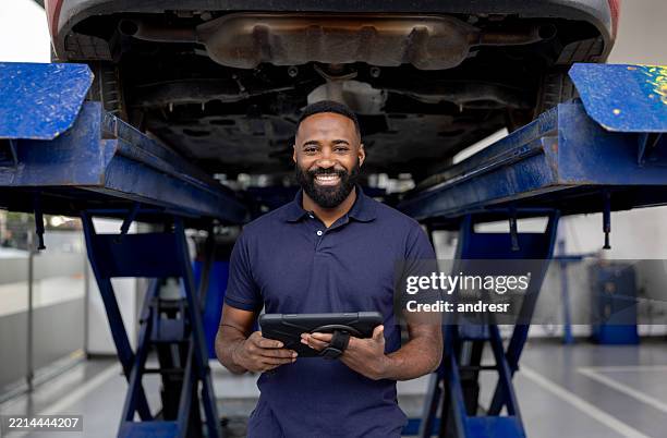 mechanic working at a car garage using a digital tablet and smiling - work overalls stock pictures, royalty-free photos & images