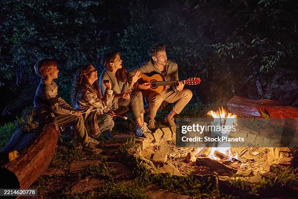 padre despreocupado tocando una guitarra para su familia junto a la hoguera por la noche. - hoguera de campamento fotografías e imágenes de stock