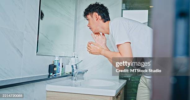 young man smiling while applying moisturizer to his face in front of mirror in the bathroom at home. healthy lifestyle in morning routine concept. - natural aging male body stock pictures, royalty-free photos & images
