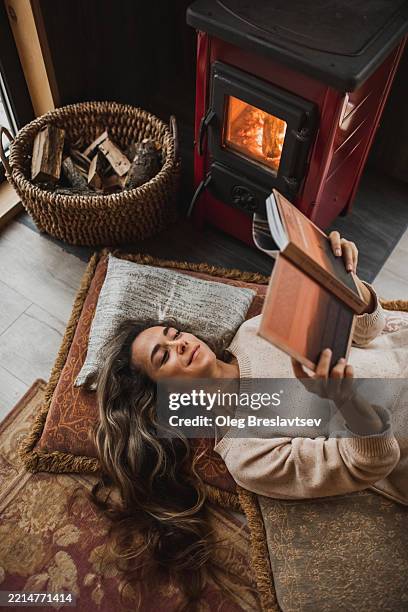 joyful woman lying on pillows near fireplace and reading a book - haardvuur stockfoto's en -beelden
