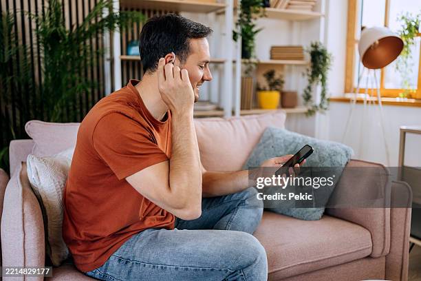 man with hearing aid using smartphone while sitting on sofa - deaf man stock pictures, royalty-free photos & images