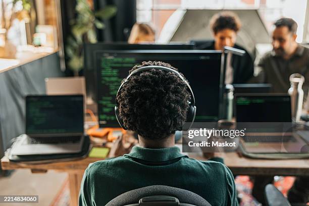 rear view of curly haired male computer programmer wearing wireless headphones coding on desktop pc while sitting on cha - programmer stock pictures, royalty-free photos & images
