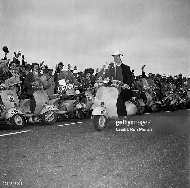 Crowd of people on Vespa mopeds waving while a vicar rides passed them on his moped during a Vespa rally, Bristol, June 22nd 1956.