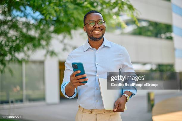 smiling businessman using smartphone and holding laptop walking outdoors - green suit stock pictures, royalty-free photos & images