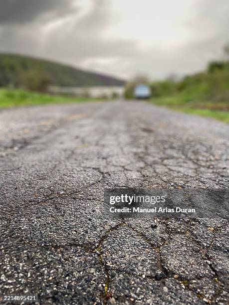 cracked asphalt road leading to a serene landscape with overcast sky and distant vehicle - condition stock pictures, royalty-free photos & images