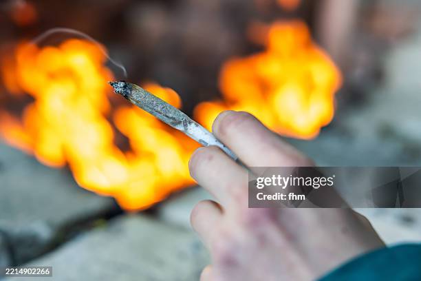 human hand holds a smoking joint with campfire in background - narcotic stock pictures, royalty-free photos & images