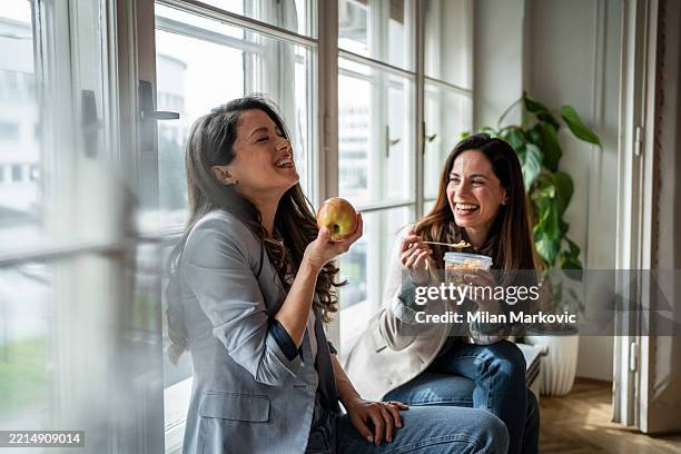 zwei fröhliche geschäftsfrauen essen gesund und lachen am fenster - ruhen stock-fotos und bilder