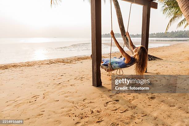 frau, die sich während des sonnenuntergangs mit palmen auf einer schaukel an einem tropischen strand entspannt - schaukel frau stock-fotos und bilder