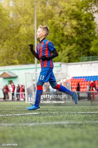 soccer player wearing number 13 jersey runs across field. teenage boy in blue and red striped uniform competes during match with spectators watching. - russian amateur football league stock pictures, royalty-free photos & images