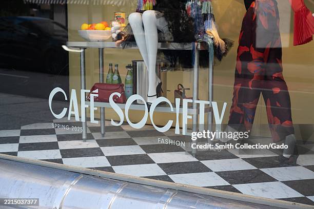 Storefront window display with CAFE SOCIETY lettering, mannequins dressed in spring fashions, tabletop fruit and beverage props, and black-and-white...