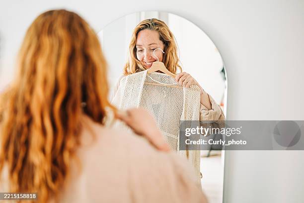 smiling woman choosing a dress in front of a mirror. - aankleden stockfoto's en -beelden