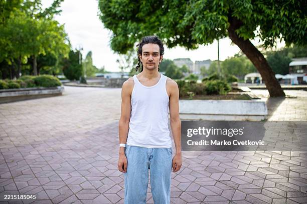 portrait of a young urban gen z man in jeans and a white sleeveless t-shirt, standing in a park and looking at the camera with dreadlocks tied back. - sleeveless stock pictures, royalty-free photos & images