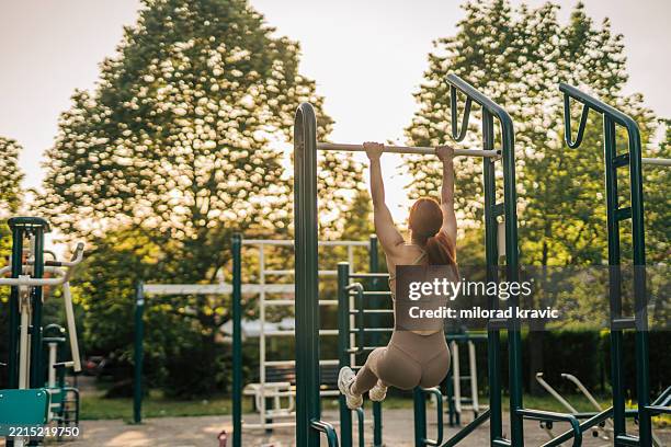 young woman performing pull-ups on a horizontal bar in an outdoor gym at sunset - horizontal bar stock pictures, royalty-free photos & images
