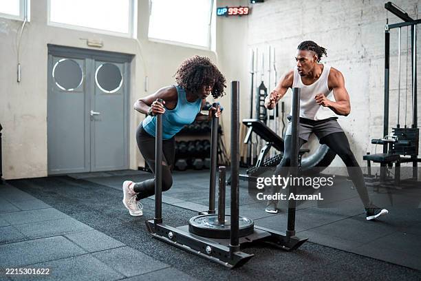focused black woman pushing sled with motivational trainer in modern gym - sled push stock pictures, royalty-free photos & images