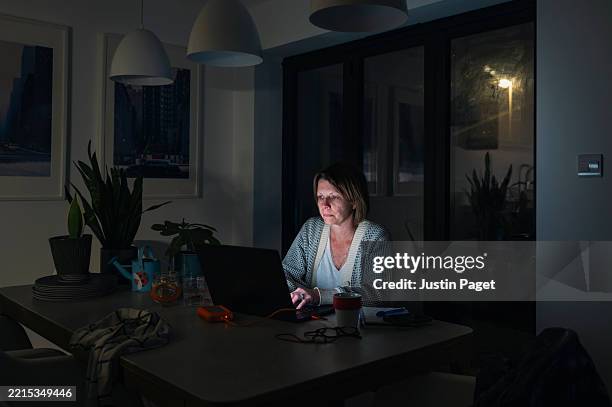 a mature woman works from home from her dining table late into the evening. she takes a break to look at her smartphone - navigare in internet foto e immagini stock