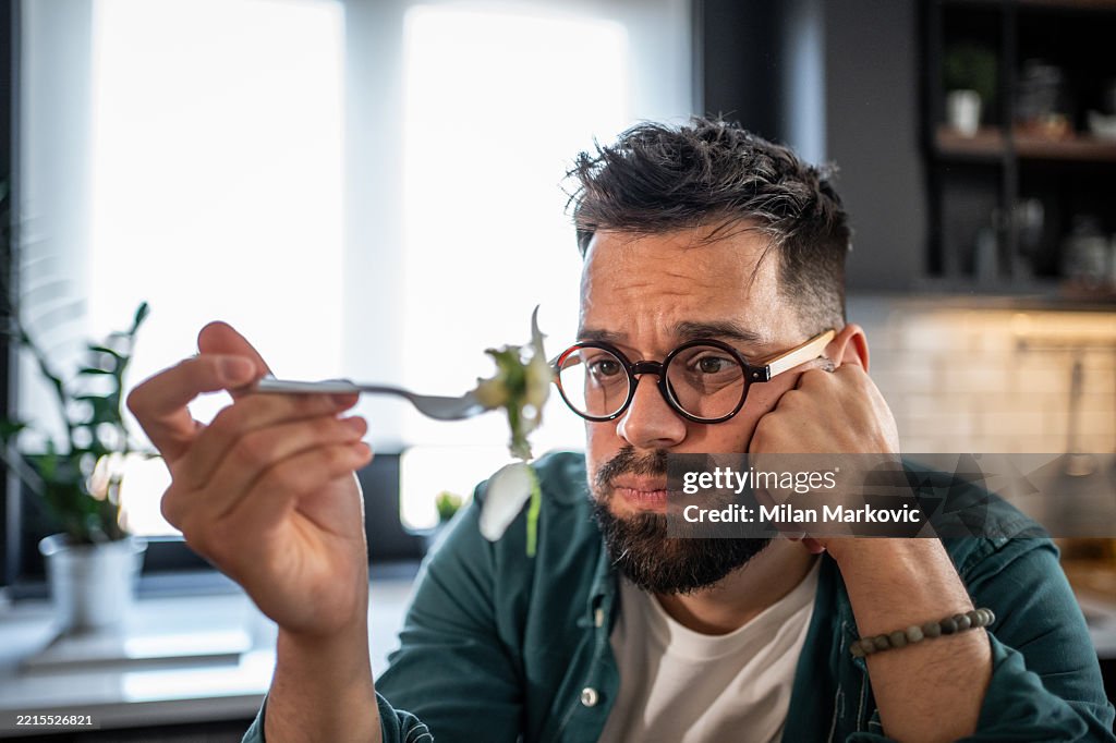 Sad young man disgusted by unhealthy food on fork in kitchen